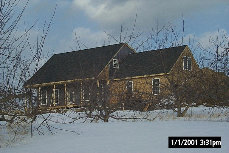 The house under construction in winter &mdash; roofed and sheathed, porch still open framing, snow to the treeline.